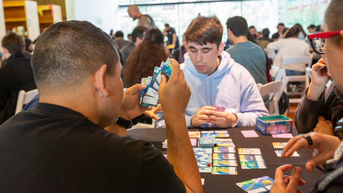 Jugadores en una mesa del Torneo ILAN en la BiblioGAM, con cartas en la mano y la sala llena de fondo